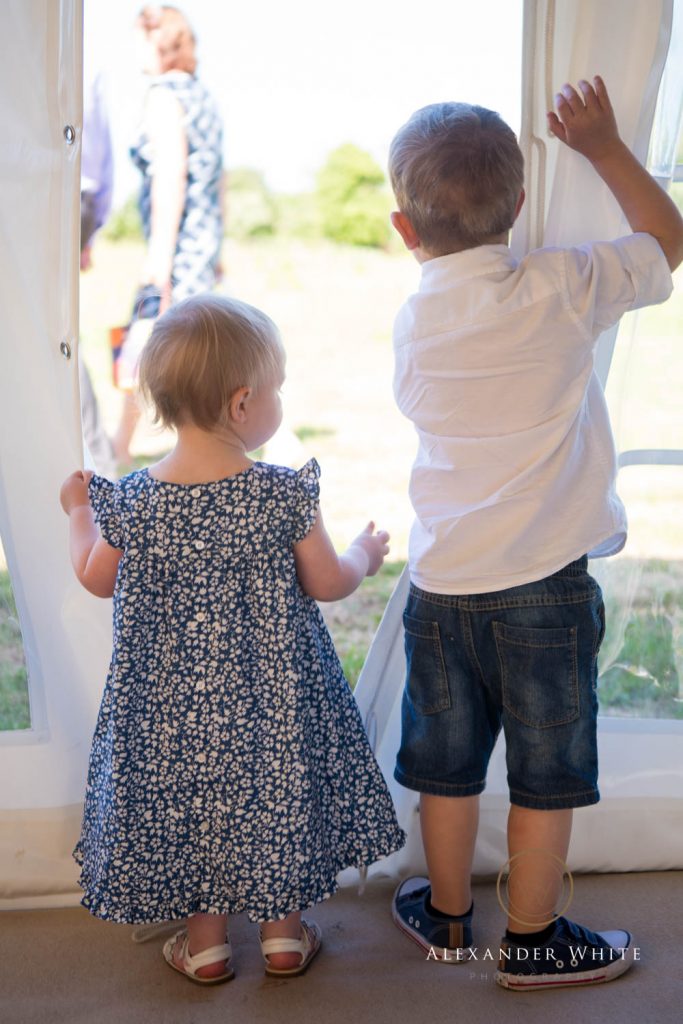 Two toddlers stand watching the goings on outside through a gap in a marquee panel.