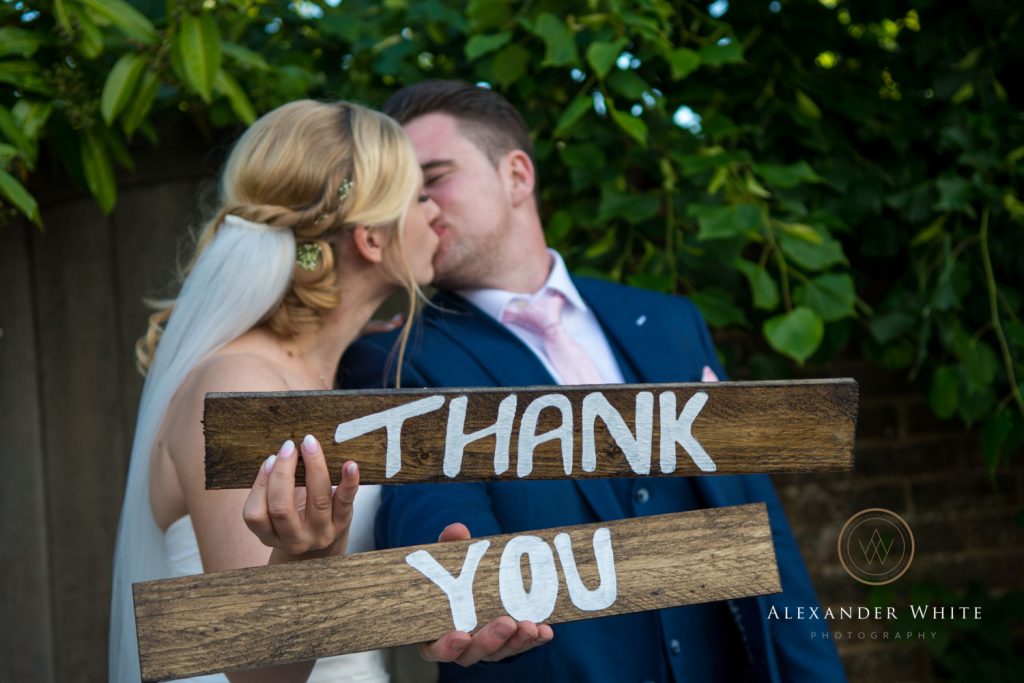 Wedding photo of the bride and groom kissing whilst holding a sign saying 'Thank You'