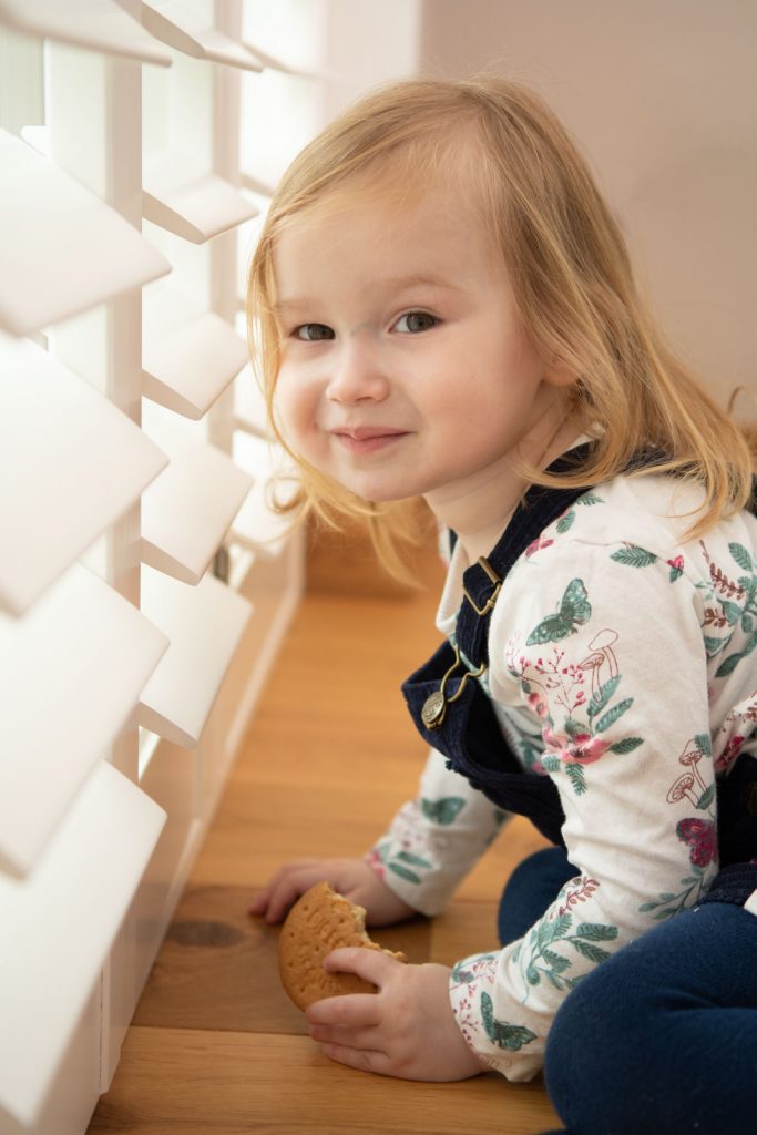 Lifestyle photography portrait of a little girl looking out of a slatted window